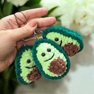 May include: Three handmade avocado keychains. Each keychain features a green crocheted exterior, a light green interior, and a brown seed. The avocados have smiling faces and are attached to silver keyrings.
