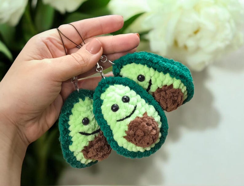 May include: Three handmade avocado keychains. Each keychain features a green crocheted exterior, a light green interior, and a brown seed. The avocados have smiling faces and are attached to silver keyrings.