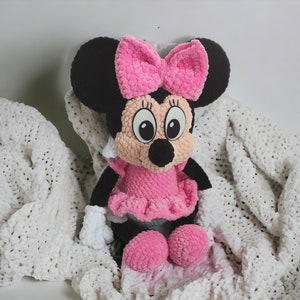 May include: A handmade Minnie Mouse plush doll. The doll has black ears, a pink bow, and a pink dress. The doll is sitting on a white blanket. The doll's face is beige with black eyes and a black nose.