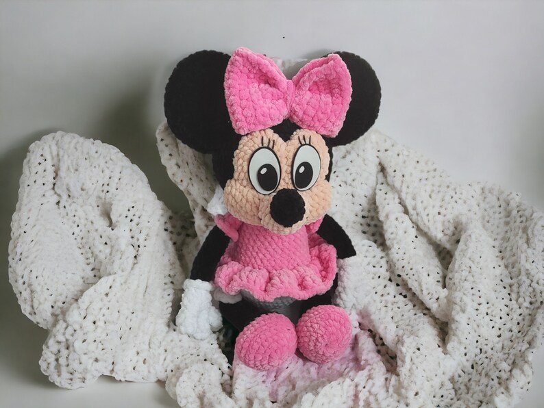 May include: A handmade Minnie Mouse plush doll. The doll has black ears, a pink bow, and a pink dress. The doll is sitting on a white blanket. The doll's face is beige with black eyes and a black nose.