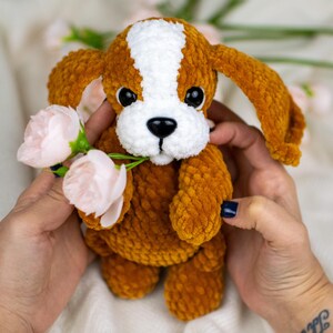 May include: A handmade crochet puppy dog with brown and white fur, black eyes, and a black nose. The plush toy is holding pink flowers. The dog is being held by a person.