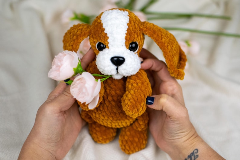 May include: A handmade crochet puppy dog with brown and white fur, black eyes, and a black nose. The plush toy is holding pink flowers. The dog is being held by a person.