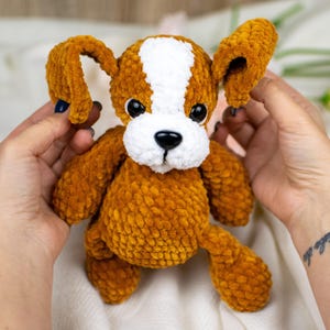 May include: A handmade, plush dog toy in shades of brown and white. The dog has large, floppy ears, a white stripe down its head, and a black nose. The toy is soft and cuddly, perfect for children or as a decorative item.