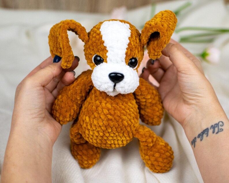 May include: A handmade, plush dog toy in shades of brown and white. The dog has large, floppy ears, a white stripe down its head, and a black nose. The toy is soft and cuddly, perfect for children or as a decorative item.
