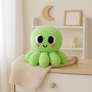 Op de afbeelding: Een limoengroen gehaakt octopus knuffel met grote witte en zwarte ogen, roze wangen en een lachende mond. De octopus zit op een crèmekleurige gebreide deken. Het speelgoed staat op een witte commode.
