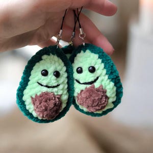 May include: Two handmade avocado-shaped keychains. Each keychain features a light green center, dark green outer edge, and a brown pit. The avocados have smiling faces with black eyes and a stitched mouth.