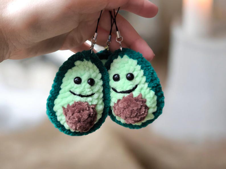 May include: Two handmade avocado-shaped keychains. Each keychain features a light green center, dark green outer edge, and a brown pit. The avocados have smiling faces with black eyes and a stitched mouth.
