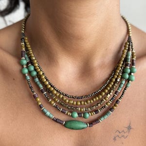 May include: A multi-strand necklace featuring a mix of beads in shades of green, brown, and gold. The necklace has a layered design with various shapes and sizes of beads, creating a bohemian style.