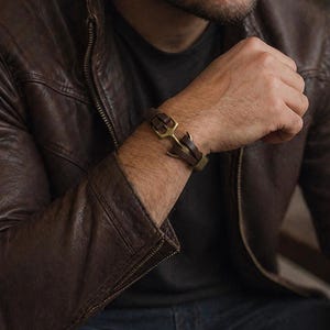 Könnte beinhalten: Ein braunes Lederarmband mit goldfarbenem Verschluss wird am Handgelenk getragen. Das Armband wird mit einer braunen Lederjacke und einer Tasse Kaffee kombiniert. Das Bild hat eine warme, einladende Ästhetik.