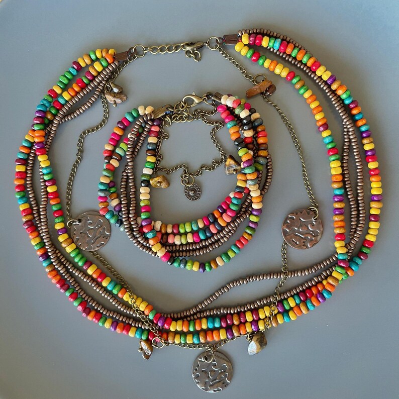 May include: A multi-strand necklace and bracelet set. The jewelry features colorful wooden beads in a rainbow of hues, strung together with metal chains and accents. The necklace has three decorative metal pendants.