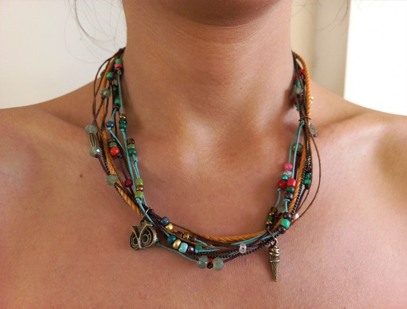 Op de afbeelding: Een meerstrengs ketting met bruine, turquoise en gouden koorden. De ketting heeft kleurrijke kralen in rood, groen en blauw, samen met kleine bedels, waaronder een uil en een ijsje. De ketting is ontworpen om om de nek te worden gedragen.
