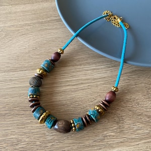 May include: A turquoise and brown beaded necklace with gold accents. The necklace features a variety of beads, including wooden, patterned ceramic, and metal beads. The necklace is strung on a turquoise cord and has a gold clasp.