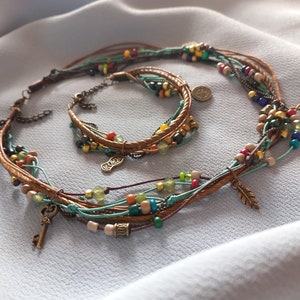 Op de afbeelding: Een meerstrengs ketting- en armbandset. De sieraden hebben bruine leren koorden, kleurrijke kralen in verschillende maten en kleuren en kleine metalen bedels. De ketting en armband worden gepresenteerd op een lichtblauwe stof.