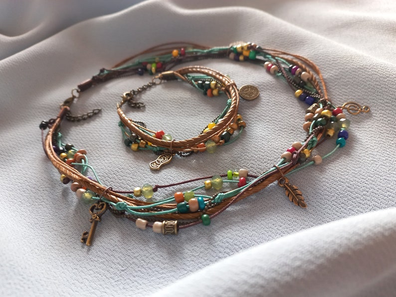Op de afbeelding: Een meerstrengs ketting- en armbandset. De sieraden hebben bruine leren koorden, kleurrijke kralen in verschillende maten en kleuren en kleine metalen bedels. De ketting en armband worden gepresenteerd op een lichtblauwe stof.