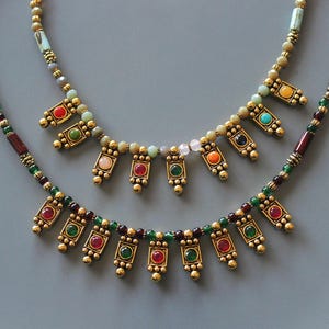 Può includere: Due collane con pendenti color oro e pietre preziose colorate. Le collane hanno un design a perline con pietre rosse, verdi, arancioni, rosa e blu. Le collane sono esposte su una superficie grigia.