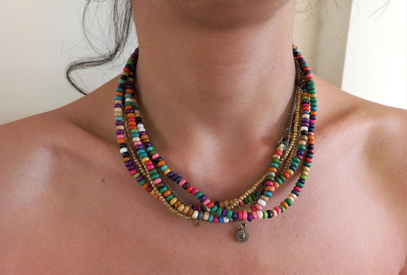 May include: A multi-strand necklace featuring colorful wooden beads in various hues, including pink, blue, green, and orange. The necklace has a small, round silver pendant and a gold-colored chain. This bohemian-style necklace is a vibrant accessory.