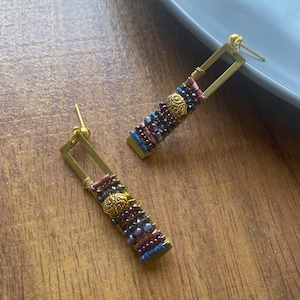 Könnte beinhalten: Ein Paar rechteckige Ohrringe mit goldfarbenen Rahmen. Die Ohrringe sind mit einer Mischung aus kleinen, farbigen Perlen in Blau-, Lila- und Rosatönen verziert. Ein goldfarbenes Dekorationselement befindet sich in der Mitte jedes Ohrrings.