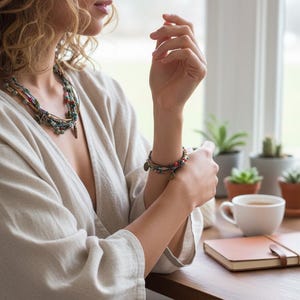 Op de afbeelding: Een vrouw draagt een beige linnen top, een ketting met meerdere strengen en een armband met kleine bedels. De sc&egrave;ne omvat een houten tafel, een kopje koffie, een notitieboek en potplanten bij een raam.