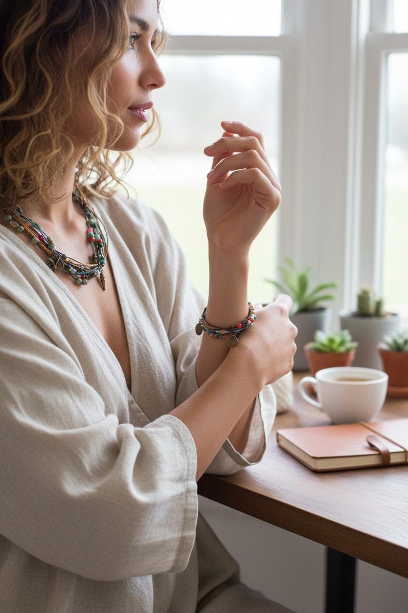 Op de afbeelding: Een vrouw draagt een beige linnen top, een ketting met meerdere strengen en een armband met kleine bedels. De sc&egrave;ne omvat een houten tafel, een kopje koffie, een notitieboek en potplanten bij een raam.