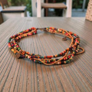 May include: A multi-strand necklace with colorful beads in shades of orange, green, purple, and brown. The necklace has a clasp and small metal charms. The necklace is displayed on a wooden surface.