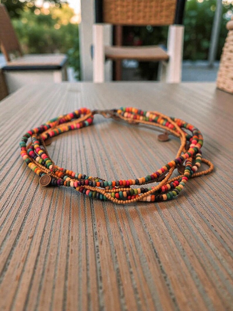 May include: A multi-strand necklace with colorful beads in shades of orange, green, purple, and brown. The necklace has a clasp and small metal charms. The necklace is displayed on a wooden surface.