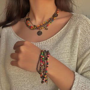 May include: A multi-strand necklace and bracelet set. The necklace features colorful beads, a circular pendant, and a heart charm. The bracelet has similar colorful beads and charms. The jewelry is displayed on a person wearing a cream-colored sweater.