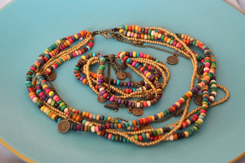 May include: A multi-strand necklace and bracelet set. The necklace and bracelet feature colorful wooden beads in various colors, including red, yellow, green, and blue. Gold-colored chains and small coin charms add detail.