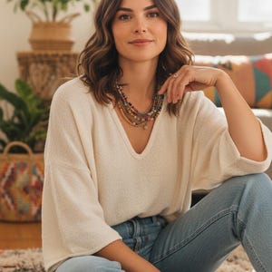 May include: A woman wearing a cream-colored, V-neck sweater and light blue jeans. She is wearing a multi-strand beaded necklace with various colors. The setting appears to be a living room with natural light.