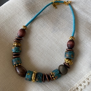 May include: A handcrafted necklace featuring a turquoise suede cord and a variety of beads. The beads include dark wood, gold-toned metal, and patterned teal and gold glass beads. The necklace is secured with a gold-toned clasp.