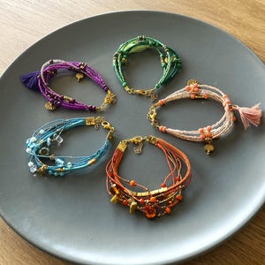 Könnte beinhalten: Fünf bunte Perlenarmbänder mit goldenen Akzenten und Quasten auf einem grauen Teller. Die Armbänder bestehen aus verschiedenen Materialien, darunter Perlen, Schnur und Leder.