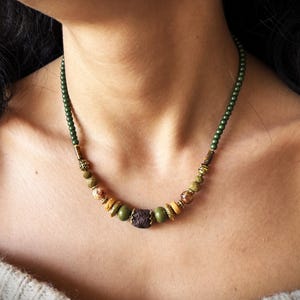 May include: A green, brown, and gold beaded necklace with a lava rock bead in the center. The necklace is on a person's neck.