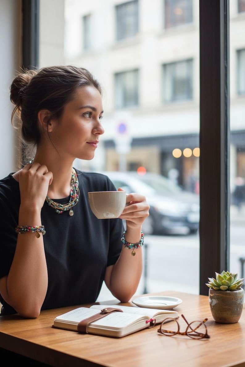 Op de afbeelding: Een persoon in een zwart topje zit aan een houten tafel en houdt een witte koffiekop vast. Ze draagt een kralenketting en armbanden. Een notitieboek, een bril en een kleine vetplant staan op tafel.