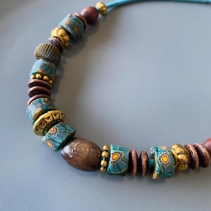 May include: A handcrafted necklace featuring a variety of beads. The necklace includes wooden, gold-toned, and turquoise-colored beads with floral patterns. The necklace is strung on a teal cord, creating a unique and artistic accessory.