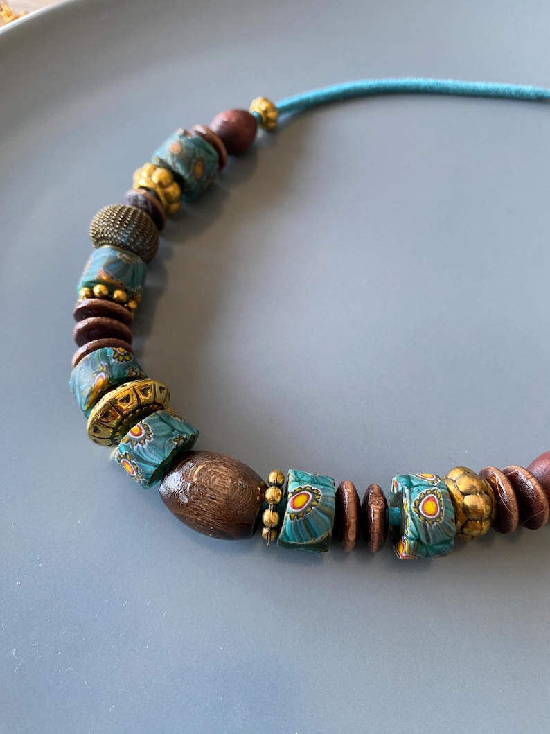 May include: A handcrafted necklace featuring a variety of beads. The necklace includes wooden, gold-toned, and turquoise-colored beads with floral patterns. The necklace is strung on a teal cord, creating a unique and artistic accessory.