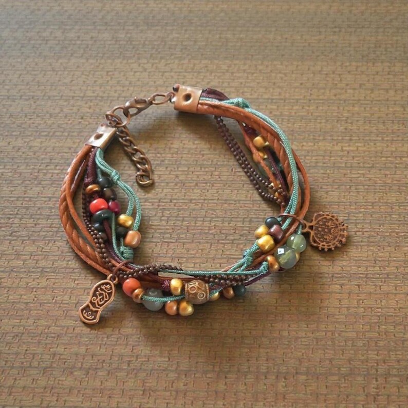Op de afbeelding: Een armband met meerdere strengen met leer, koord en kettingschakels in bruin, blauwgroen en bordeaux. De armband is versierd met kralen in verschillende kleuren en decoratieve metalen bedels. De sluiting is koperkleurig.