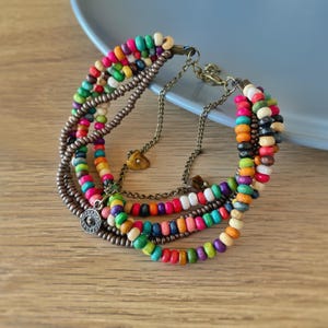 May include: A multi-strand bracelet featuring colorful wooden beads in various hues, including red, green, orange, and blue. The bracelet has a bronze-colored chain and clasp, with decorative charms.