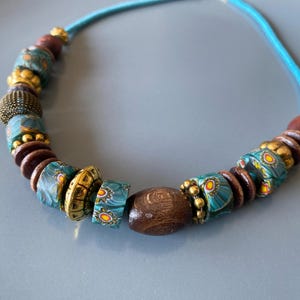 May include: A handcrafted necklace featuring a turquoise cord strung with a variety of beads. The beads include dark brown wooden beads, gold-colored metal beads, and patterned turquoise and yellow glass beads. The necklace is displayed on a gray surface.