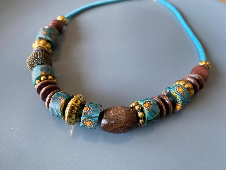 May include: A handcrafted necklace featuring a turquoise cord strung with a variety of beads. The beads include dark brown wooden beads, gold-colored metal beads, and patterned turquoise and yellow glass beads. The necklace is displayed on a gray surface.