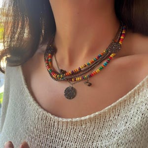 May include: A multi-strand necklace featuring colorful wooden beads in a rainbow pattern. The necklace has metal chain accents and decorative coin pendants. The necklace is a bohemian style.