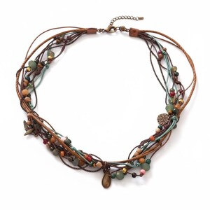 Op de afbeelding: Een meerstrengs ketting met bruine leren koorden en kralen in groen, oranje en bruin. De ketting heeft kleine metalen bedels en een sluiting. De ketting is ongeveer 40 cm lang.