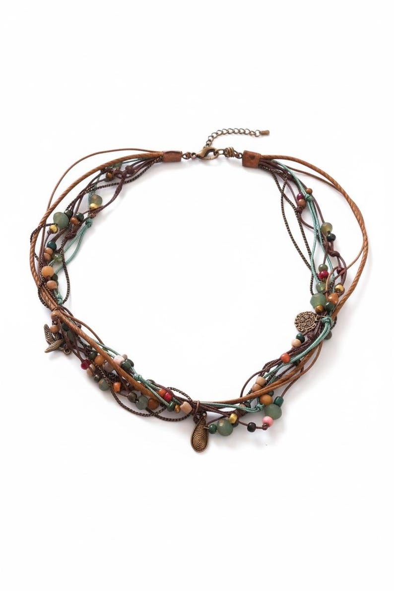 Op de afbeelding: Een meerstrengs ketting met bruine leren koorden en kralen in groen, oranje en bruin. De ketting heeft kleine metalen bedels en een sluiting. De ketting is ongeveer 40 cm lang.