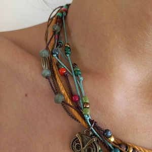 Op de afbeelding: Een meerstrengs ketting met kleurrijke kralen en een bronzen uil hanger. De ketting heeft strengen van bruine, blauwgroene en paarse koorden, geaccentueerd met groene, rode en blauwe kralen. De uil hanger voegt een grillige toets toe.