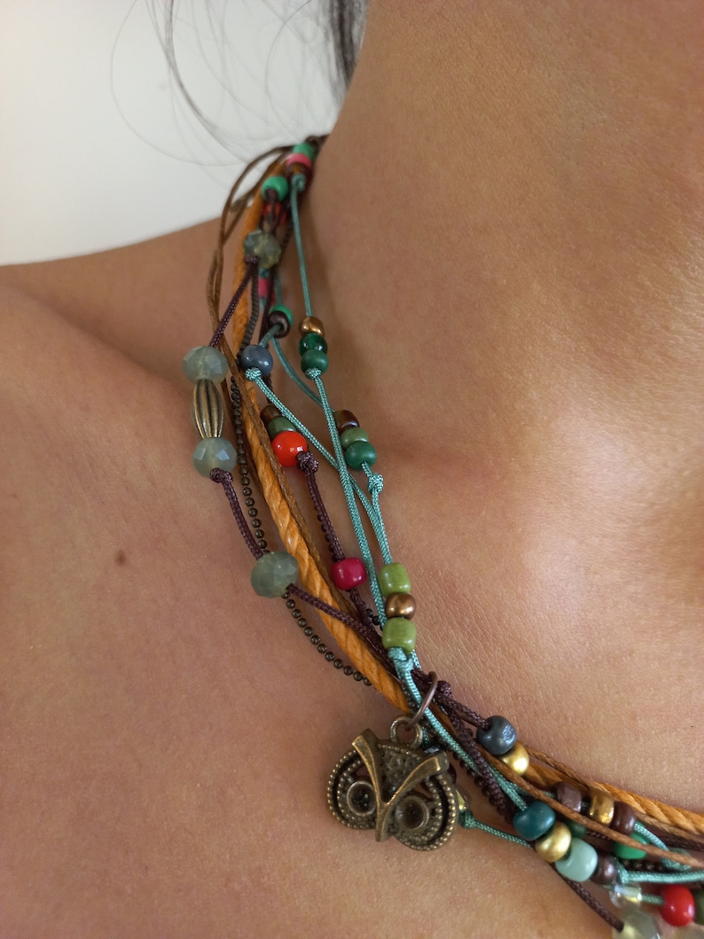 Op de afbeelding: Een meerstrengs ketting met kleurrijke kralen en een bronzen uil hanger. De ketting heeft strengen van bruine, blauwgroene en paarse koorden, geaccentueerd met groene, rode en blauwe kralen. De uil hanger voegt een grillige toets toe.