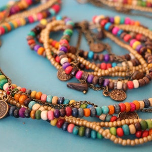 May include: A collection of colorful beaded necklaces and bracelets. The necklaces feature wooden beads in various colors, including red, yellow, green, and blue, with small metal coin charms. The bracelets are made of smaller, gold-colored beads.