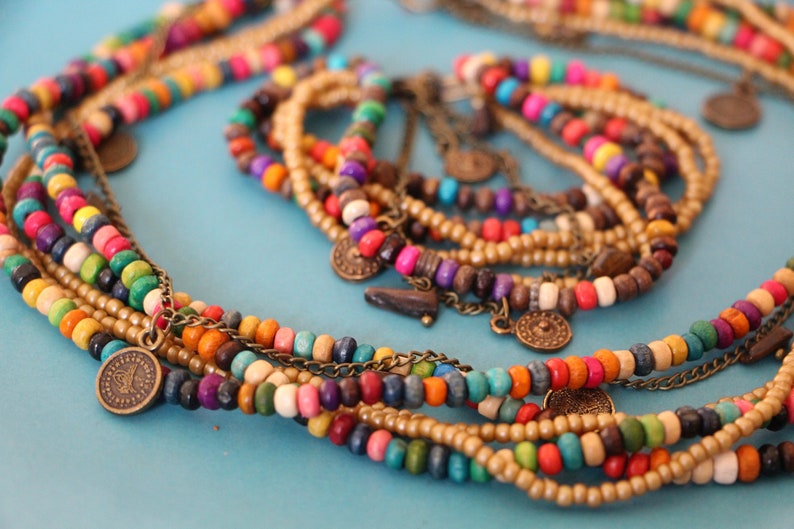 May include: A collection of colorful beaded necklaces and bracelets. The necklaces feature wooden beads in various colors, including red, yellow, green, and blue, with small metal coin charms. The bracelets are made of smaller, gold-colored beads.
