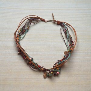 Op de afbeelding: Een meerstrengs ketting met bruine leren koorden, turquoise, bruine en rode draden en diverse kleurrijke kralen. De ketting heeft een druppelvormige hanger en een kleine bedel. De sieraden worden gepresenteerd op een licht houten oppervlak.