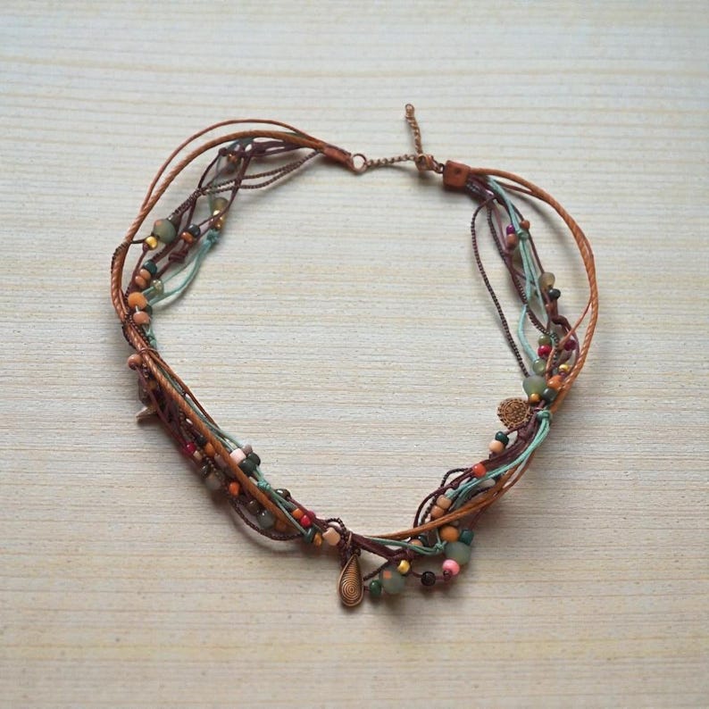 Op de afbeelding: Een meerstrengs ketting met bruine leren koorden, turquoise, bruine en rode draden en diverse kleurrijke kralen. De ketting heeft een druppelvormige hanger en een kleine bedel. De sieraden worden gepresenteerd op een licht houten oppervlak.