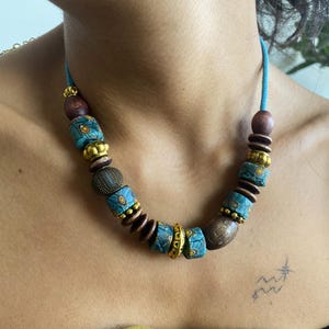 May include: A turquoise beaded necklace with a variety of beads. The necklace features wooden, gold, and patterned turquoise beads. The necklace is strung on a turquoise cord. The necklace is a unique piece of jewelry.