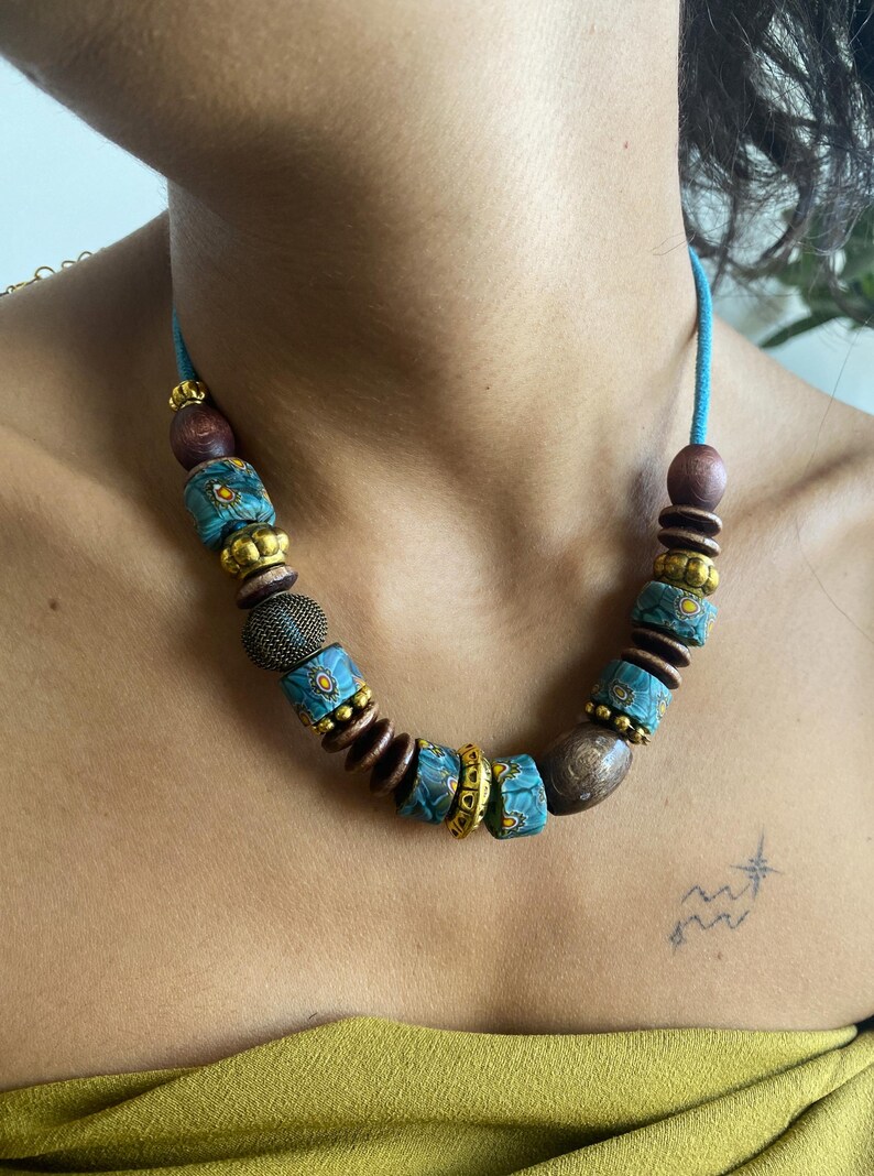 May include: A turquoise beaded necklace with a variety of beads. The necklace features wooden, gold, and patterned turquoise beads. The necklace is strung on a turquoise cord. The necklace is a unique piece of jewelry.
