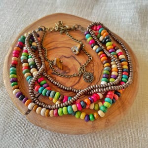 May include: A collection of colorful beaded necklaces and a chain necklace with decorative charms. The necklaces feature wooden beads in various colors, including red, orange, yellow, green, blue, and purple. The necklaces are arranged on a wooden surface.